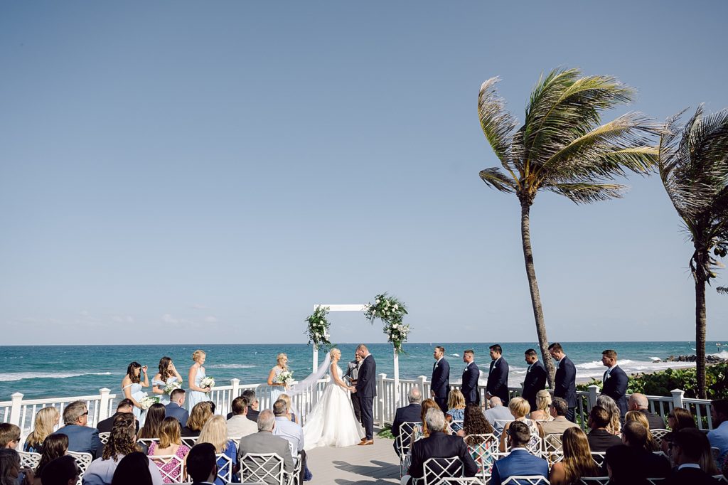 Wide shot of Bride and Groom hand in hand for outdoor beach Ceremony with Ocean in front of Guests